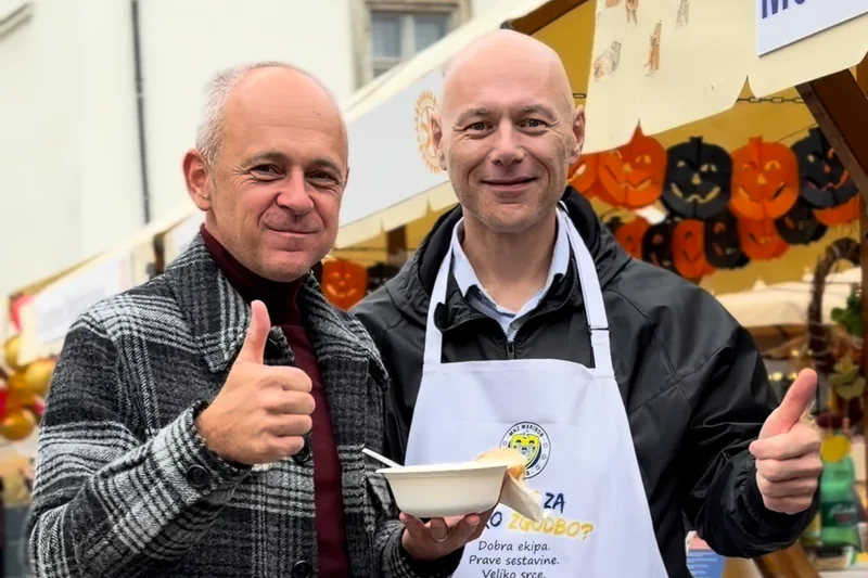 Dva udeleženca Rotarijskega piskra ob stojnici, s predpasnikom in posodo jedi na žlico.